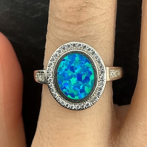May include: Silver ring with an oval blue-green opal stone. The opal is encircled by a halo of small, clear gemstones. The ring band has clear gemstones on either side of the opal.