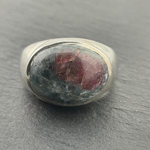 May include: A silver ring with a large oval-shaped gemstone. The gemstone is a mix of green, gray, and red.