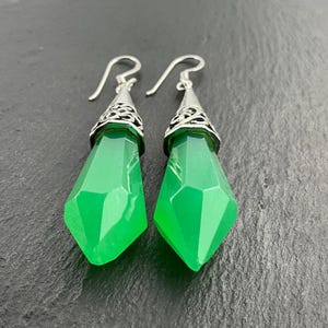 May include: A pair of green gemstone earrings with silver-colored hooks. The earrings feature a faceted, teardrop-shaped green stone with a silver filigree design at the top. The earrings are displayed against a dark background.