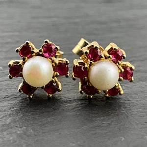 May include: A pair of gold stud earrings, each featuring a central round white pearl surrounded by six small, vibrant red rubies arranged in a floral pattern. The metal settings are yellow gold, and the design creates a classic, symmetrical look.