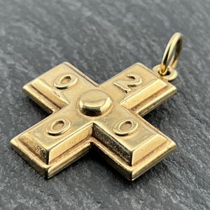 Puede incluir: Colgante de cruz de color dorado con un lazo para una cadena. La cruz tiene un diseño en relieve con un círculo central y cuatro círculos más pequeños. El número "12" es visible en un brazo.