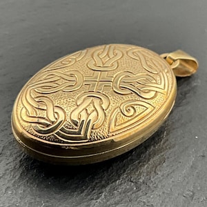 May include: An oval gold-toned locket pendant featuring an intricate raised knotwork pattern across the front surface. The metal has a textured background with polished, smooth raised lines forming the design, and includes a small top bail.