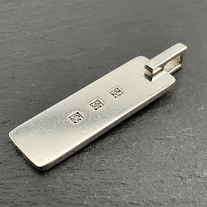 May include: A silver pendant featuring three small square gemstones. The pendant has a rectangular design with a loop for a chain. The gemstones are evenly spaced along the length of the pendant. A modern piece of jewellery.