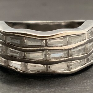 Puede incluir: Un anillo de plata con una banda ondulada y cuatro filas de piedras talla baguette transparentes.