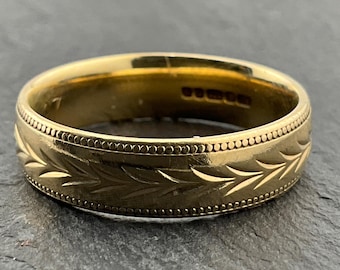 May include: A gold ring featuring an engraved leaf design. The ring has a beaded edge and a smooth inner surface. The ring is displayed on a dark grey surface.