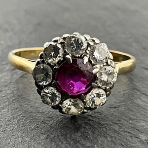 May include: A gold ring with a central, round, deep pink ruby encircled by a halo of clear, round diamonds. The ring's band is plain gold, and the setting combines gold and silver. A classic, elegant design.