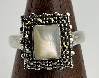 May include: A silver ring with a square, mother-of-pearl center stone. The ring is surrounded by a square frame with small, dark, round accents. The ring is displayed on a wooden stand.