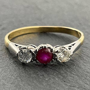 May include: A three-stone ring featuring a central ruby gemstone flanked by two clear diamonds. The ring has a gold band with silver accents near the gemstones. The gemstones are set in a decorative setting.