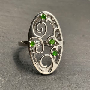 May include: A silver ring with an oval-shaped design featuring a delicate, intricate filigree pattern. Four small green gemstones are set within the design.