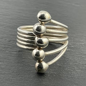 Puede incluir: Un anillo plateado con un diseño en espiral. El anillo presenta múltiples bandas que se enrollan alrededor del dedo, con cuatro acentos esféricos. El anillo está hecho de un metal brillante y reflectante.