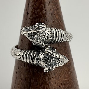 Peut inclure: Une bague en argent avec deux têtes de serpent entrelacées. La bague est détaillée avec des écailles texturées et une bande de lignes en relief.