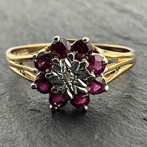 May include: A gold ring featuring a cluster of eight round, deep pink rubies arranged in a floral pattern around a central white metal setting that holds a single small, clear, round-cut diamond at its centre, with a split-shank gold band.