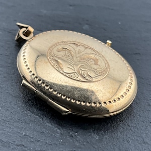 Könnte beinhalten: Ein ovaler Medaillon-Anhänger aus goldfarbenem Metall mit einem zentralen, gravierten Blumenmotiv, das von einem Perlenrand umgeben ist. Das Medaillon verfügt über einen seitlichen Scharniermechanismus und eine Öse zur Befestigung mit polierter Oberfläche.