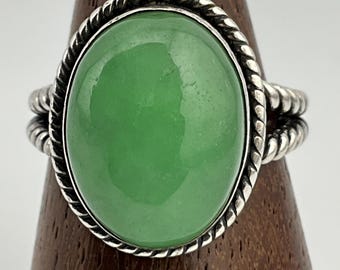 May include: Oval-shaped ring featuring a large, smooth, light green stone set in a silver-toned metal band. The band has a twisted rope design around the stone and on the sides, creating a vintage or antique aesthetic. The ring is displayed on a wooden ring holder.