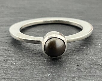 May include: A silver ring with a dark grey pearl set in the centre. The ring is a simple band with a wide, flat surface.