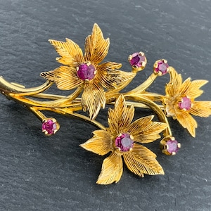Puede incluir: Un broche de tono dorado con un diseño floral estilizado con piedras preciosas rosas. El broche está hecho de metal y tiene un diseño delicado e intrincado.