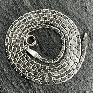 Puede incluir: Un collar de cadena de metal plateado que presenta una serie de eslabones rectangulares planos con bordes redondeados. El collar incluye un cierre de resorte circular y una lengüeta final cónica, ambos visibles en la disposición en espiral.