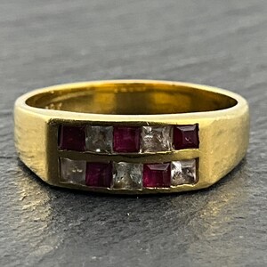 May include: A gold band ring featuring two horizontal rows of alternating square-cut red rubies and clear white diamonds. The gemstones are set in a channel setting across the front of the polished yellow gold metal band.