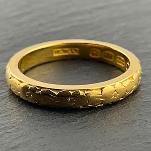 Puede incluir: Un anillo de banda de oro con un patrón floral grabado de forma continua en la superficie exterior. El interior de la banda muestra varios sellos y marcas pequeñas estampadas que indican la pureza del metal y el origen de la pieza.