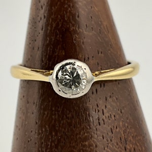 Puede incluir: Un anillo de compromiso de oro y blanco con un diamante redondo engastado en bisel.