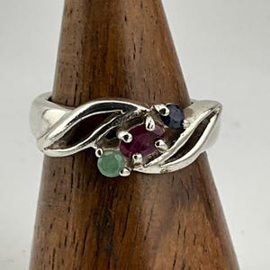 以下が含まれることがあります： ルビー、サファイア、エメラルドの3つの宝石が付いたシルバーリング。リングはねじれたバンドデザインです。宝石はクラスター状にセットされており、ユニークで人目を引くジュエリーです。