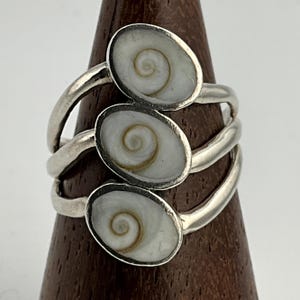 Pode incluir: Um anel de prata com três incrustações ovais de concha branca e bege. A banda do anel tem um design em camadas, com cada incrustação colocada em um nível separado. O anel é exibido em um suporte de madeira.