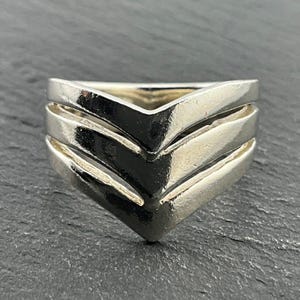 Puede incluir: Un anillo de plata en forma de chevron con tres bandas. El anillo está hecho de metal pulido y tiene un acabado liso y brillante.