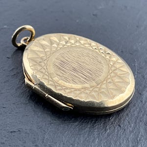 May include: An oval gold-toned locket pendant featuring a central brushed metal finish surrounded by a geometric, diamond-patterned border. The locket includes a small circular bail at the top for attachment to a chain, showcasing a hinged side design.