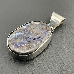 May include: A silver pendant featuring a large, oval-shaped opal. The opal displays a mix of dark gray, blue, and gold colors. The pendant has a simple, polished silver frame and a loop for a chain.