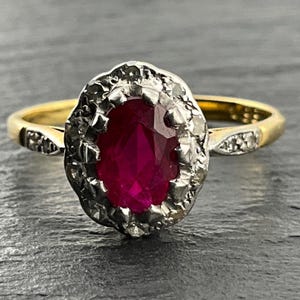 Könnte beinhalten: Ein Goldring mit einem zentralen, oval geschliffenen, tiefroten Edelstein, der von einem Kranz aus kleinen, klaren, facettierten Steinen in einer silberfarbenen Fassung umgeben ist. Der Ring hat eine glatte Goldschiene mit Verzierungen.