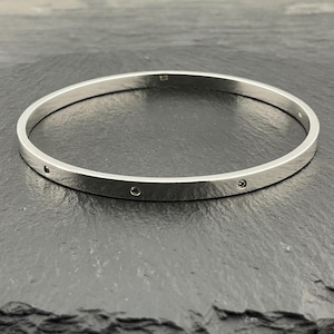 May include: A silver bangle bracelet with a flat, polished surface. The bracelet has small, evenly spaced, circular indentations. The jewelry is displayed on a dark, textured surface.