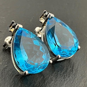 May include: A pair of silver-toned stud earrings featuring large, teardrop-shaped, bright blue gemstones. The earrings have a simple, elegant design with a small decorative element at the top of each stud. The gemstones are faceted, catching the light.