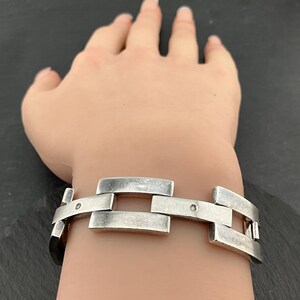 Op de afbeelding: Een zilverkleurige armband met een rechthoekig schakelontwerp. De armband, gedragen om de pols, heeft een geborstelde metalen afwerking. Kleine, ronde klinknagels verbinden de schakels. Het is een sieraad.