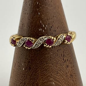 Puede incluir: Un anillo de oro con un diseño retorcido, con cinco piedras preciosas redondas de color rojo intenso y pequeñas piedras de acento transparentes. El anillo se exhibe sobre un soporte de madera, mostrando sus intrincados detalles.