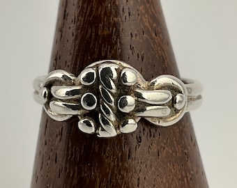 Könnte beinhalten: Ein silberner Ring mit einem detaillierten, komplizierten Design, das ein gedrehtes Seilmotiv und kleine, runde Akzente aufweist. Der Ring wird auf einem hölzernen Ringständer präsentiert.