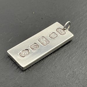 Peut inclure: Pendentif rectangulaire en argent avec une petite boucle en haut. Le pendentif comporte plusieurs poinçons estampés, dont un lion et les initiales "RJ". Le pendentif est en métal brillant et réfléchissant.