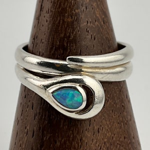 May include: A silver ring in a snake design, coiled around itself. The ring has a teardrop-shaped opal with blue and green colours. The ring is displayed on a wooden stand.