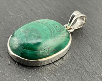 May include: A green malachite gemstone pendant set in a silver oval frame. The pendant has a bail for attaching to a chain.