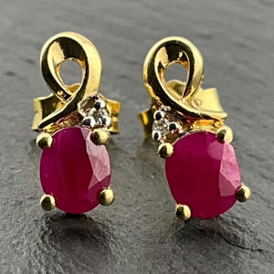 May include: A pair of gold stud earrings featuring oval-cut vibrant red rubies held by four prongs. Above each ruby is a swirling gold ribbon design accented with three small, clear, round-cut diamonds set in a white metal base.