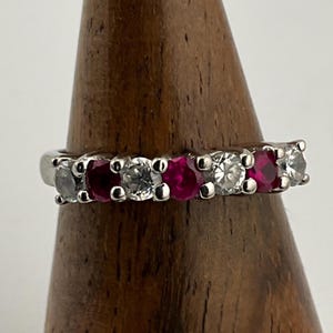May include: A silver ring featuring alternating red and clear gemstones. The ring has a row of five stones, with the red stones appearing to be rubies. The ring is displayed on a wooden stand.