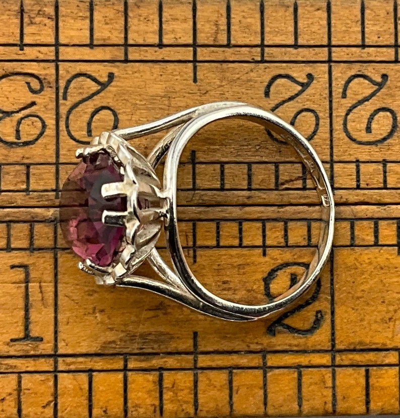 May include: A silver ring with a large, round, purple gemstone set in a prong setting. The ring is on a wooden ruler.