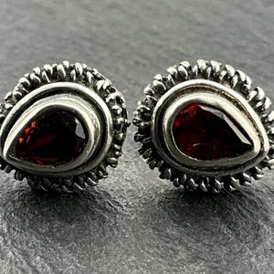 May include: A pair of silver earrings with red gemstone teardrop-shaped stones set in a textured silver frame.