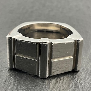 May include: A wide silver-toned metal ring featuring a geometric design with a central vertical bar flanked by two rectangular panels on each side. The band has a smooth, polished finish and a substantial, thick construction with clean, straight edges.