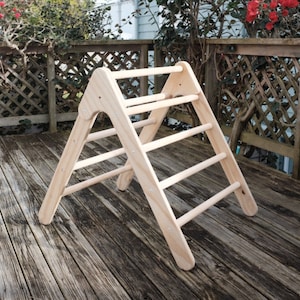 Puede incluir: Un triángulo de escalada de madera con cinco peldaños. El triángulo está hecho de madera natural y está de pie en una plataforma de madera.