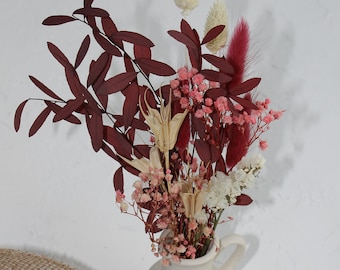 May include: A white ceramic pitcher with a bouquet of dried flowers, including pink, red, and white flowers, and brown branches. The pitcher is sitting on a brown burlap surface.