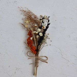 May include: A small bouquet of dried flowers and grasses featuring burnt orange, black, and white elements. The stems are bundled together and secured with a natural jute twine tied in a bow, creating a compact arrangement of dried botanical stems.