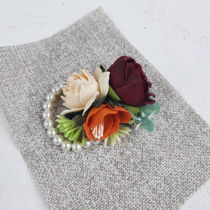 May include: A pearl bracelet with a floral arrangement featuring a burgundy, cream, and orange flower.