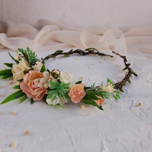 Peut inclure: Une couronne de fleurs composée d'une base en vigne tressée marron foncé, ornée de fleurs pêche, crème et blanches, de feuilles vertes et de petits bourgeons blancs. L'accessoire comprend de longs rubans beiges transparents aux extrémités.