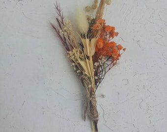 Puede incluir: Un pequeño ramo de flores secas, con tonos naranja, beige y marrón. El arreglo está atado con hilo y asegurado con un alfiler. El fondo es una superficie blanca texturizada.