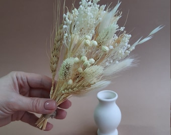 May include: A small bouquet of dried flowers and grasses in shades of cream, beige, and white, featuring wheat stalks, fluffy bunny tails, and delicate baby's breath, positioned next to a small, smooth, matte white ceramic bud vase.